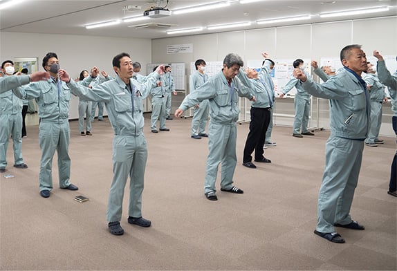 大会議室で社員全員が等間隔に並びラジオ体操を行っている様子